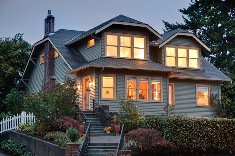 Warm, orange light glows from within the Greenlake Guest House with light blue siding and green foliage on the exterior.