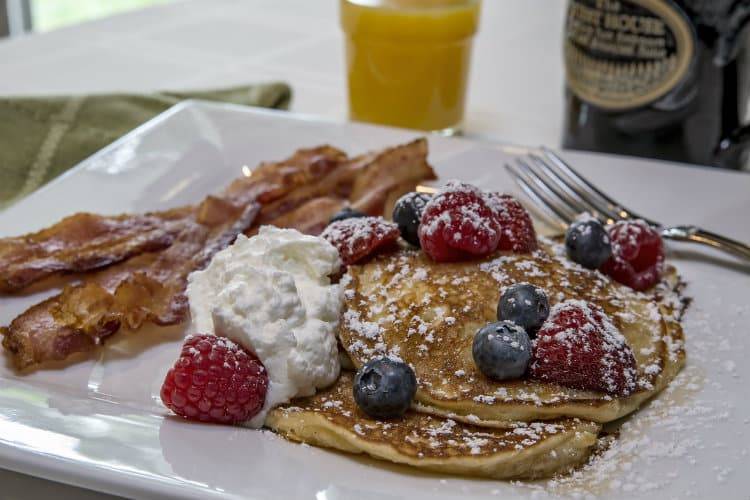 A white plate of pancakes, red raspberries, blueberries, bacon, and white cream at the Guest House B&B.