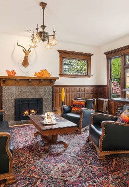 Brightly lit carpeted room with fireplace, and various cushioned chairs, windows with oak framing, rectangular coffee table