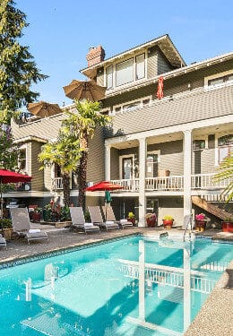 Pool with reflection of 3-story beige house, concrete patio deck with chaise loungers, tables with umbrellas