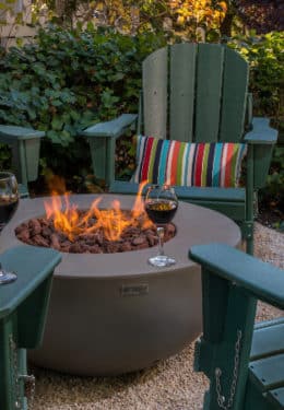 burning fire pit on concrete pad surrounded by green chairs with glasses filled with red wine