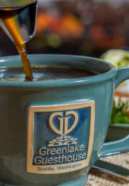 coffee being poured from stainless steel pot into green mug with Greenlake Guesthouse symbol next to breakfast plate on tan placemat