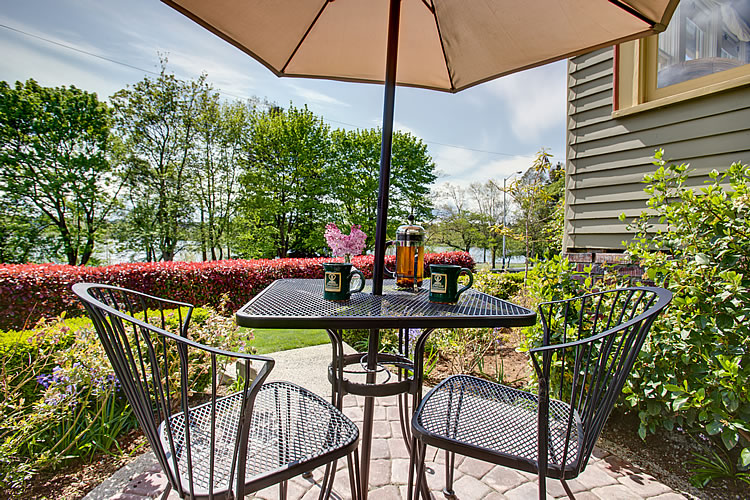 A black metal table and chairs sits under a brown umbrella amidst green foliage at the Greenlake Guest House.