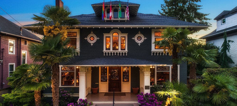 Brighly lit 3-story gray home surrounded by palm trees with 4 flags hanging, covered porch