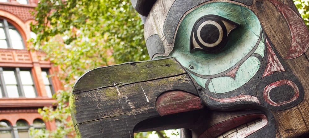 Wooden sculpture in the foreground with with brick buildings and green foliage in the background.