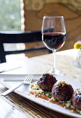 A glass of red wine sits behind a white platter holding meatballs over sauce and a fork.