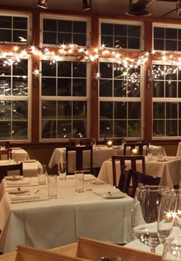 A table for four with a white tablecloth in a room with large windows and white, decorative lights.