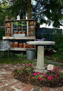 Grey stone bird bath surrounded by pink flowers and wooden shelves behind it with teracotta pots filled with white flowers.