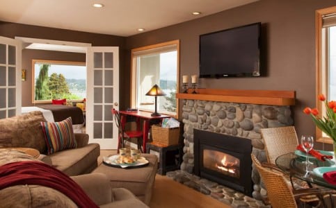 A living room with beige couches and wall with TV mounted over a stone hearthed fireplace at the Three Tree Point B&B.